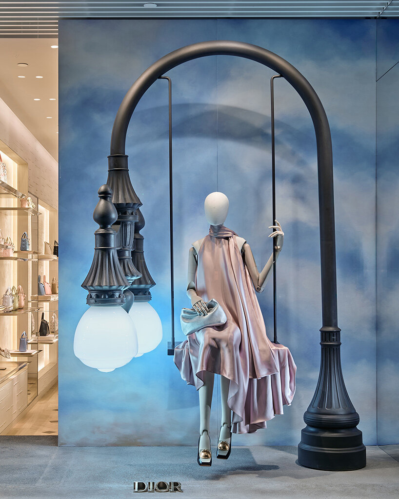 Манекен на качелях под изогнутым фонарём в витрине Dior